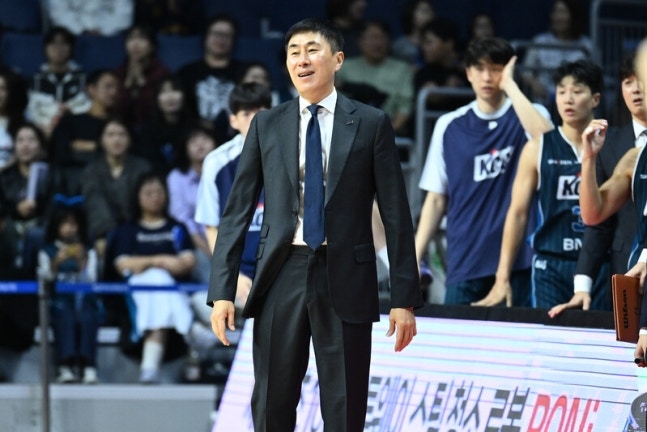 짙은 회색 정장에 푸른 넥타이를 맨 부산 KCC 이상민 감독이 코트 옆에서 웃고 있고, 뒤쪽 벤치 앞에 KCC 선수들이 서 있다