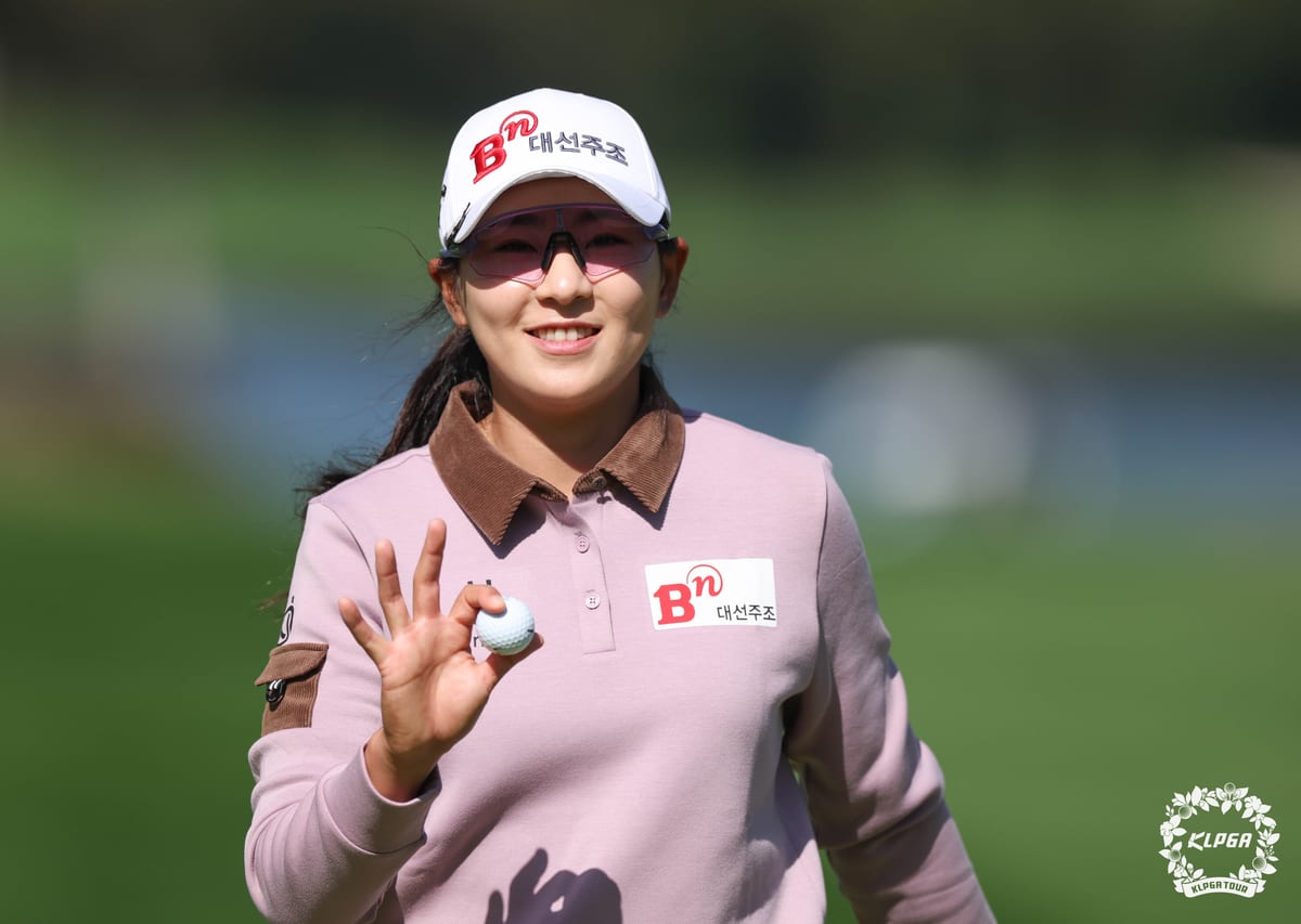 분홍 상의와 흰 모자를 쓴 여자 선수가 웃으며 오른손에 든 골프공을 들어 보이고 있다. 뒤로 초록 잔디와 물이 흐릿하게 보인다